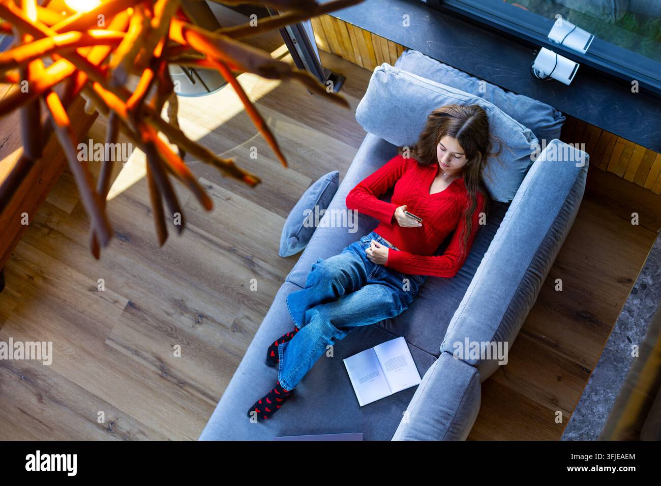 Junge Frau entspannt sich auf dem Sofa und nutzt ihr Smartphone. Junge Frau in einem roten Pullover lehnt sich auf einem grauen Sofa zurück und verwendet ihr Smartphone, mit einem offenen Buch daneben Stockfoto