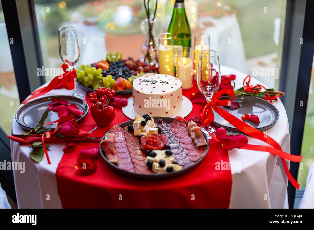 Romantischer Heiratsantragstisch mit Kuchen und Kerzen. Romantischer Antragstisch mit Heiratsantragskuchen, Kerzen, Aufschnitt, Obst, roten Bändern und Stockfoto
