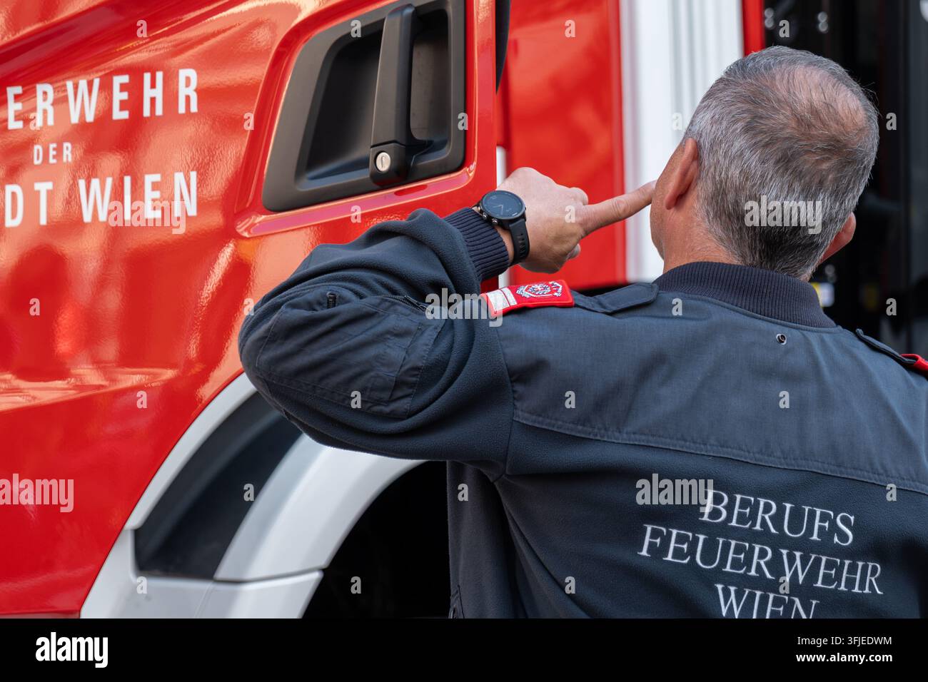 WIEN, ÖSTERREICH, 12. September 2026: Ein Feuerwehrmann zeigt auf sein Ohr, während er neben einem roten Wiener Feuerwehrwagen steht. Stockfoto