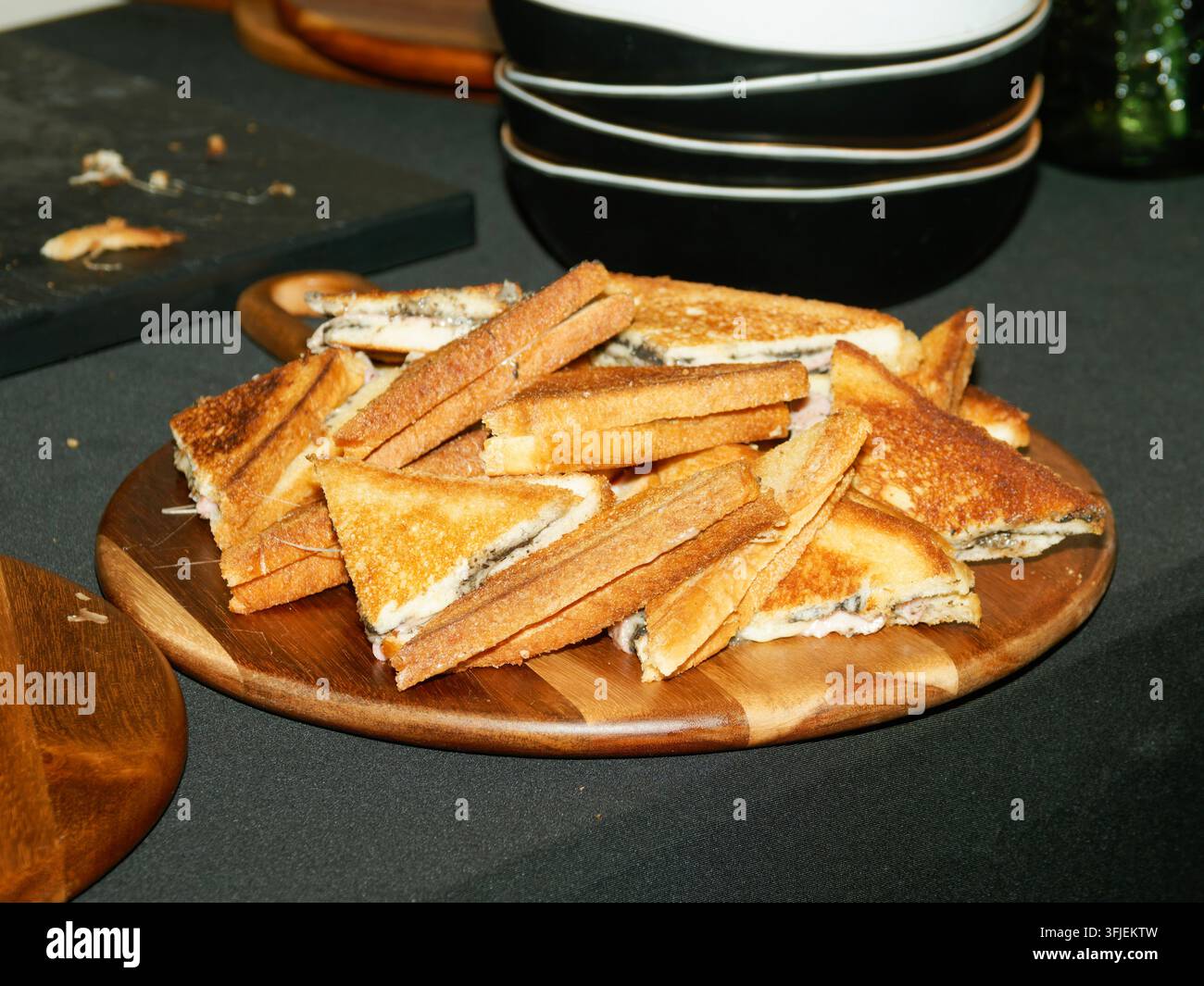 Teller mit gerösteten dreieckigen Sandwiches auf einem Holzbrett mit gestapelten Schüsseln im Hintergrund. Stockfoto