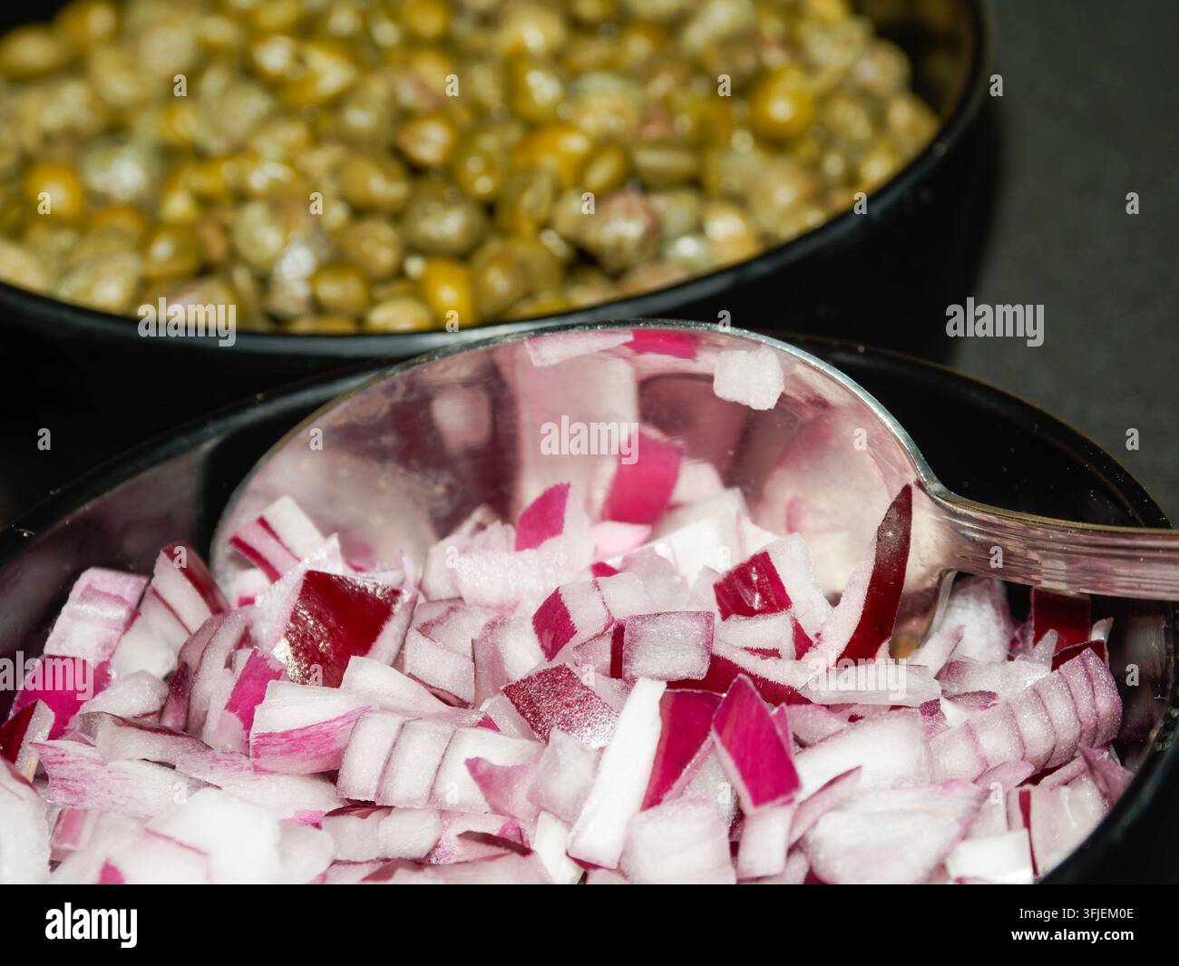 Nahaufnahme von gehackten roten Zwiebeln und grünen Erbsen in schwarzen Schüsseln mit einem Metalllöffel. Stockfoto