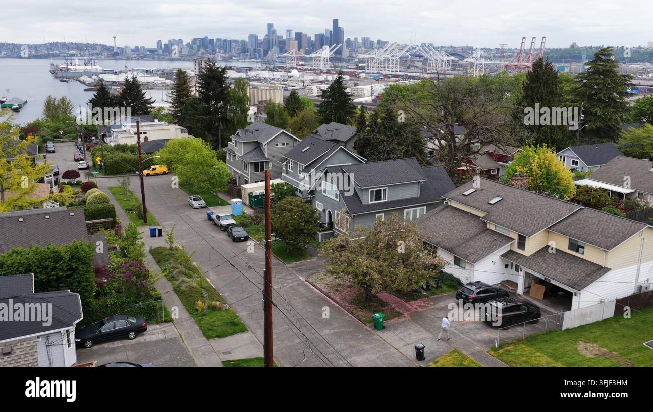 An einer hügeligen Wohnstraße mit Blick auf die Bucht und den Industriehafen erhebt sich der Space Needle und die Innenstadt-Skyline über Dächern und beschatteten Einfahrten. Seattl Stockfoto