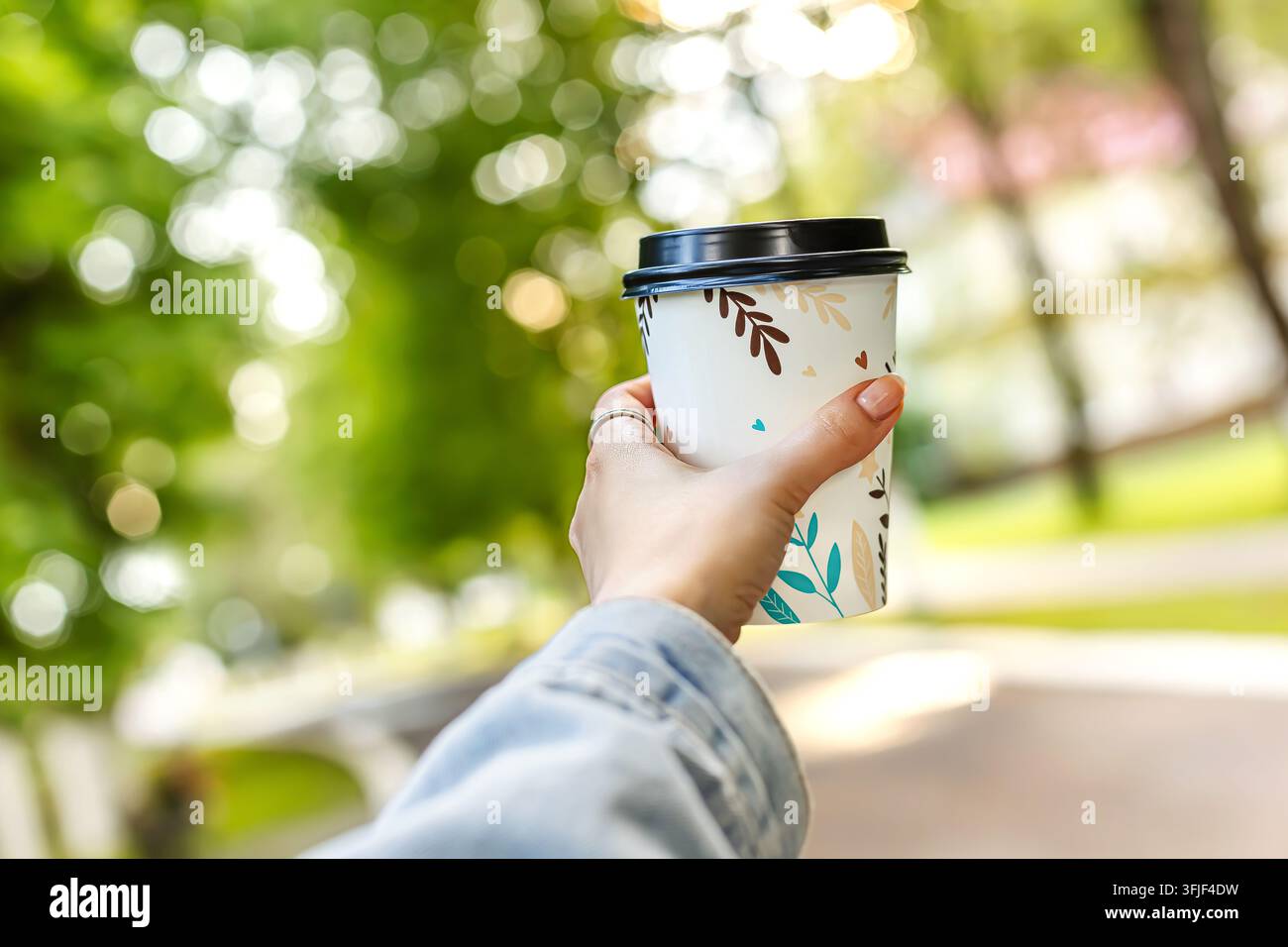Nahaufnahme einer Frauenhand, die draußen eine Papierkaffeetasse mit schwarzem Deckel hält. Heißes Getränk zum Mitnehmen in einem wunderschönen Sommerpark mit grünen Bäumen und s Stockfoto