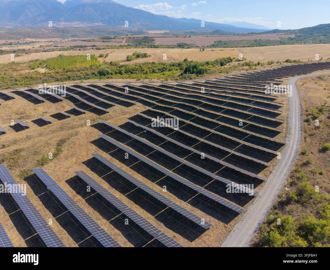 Solarmodule bilden lange parallele Reihen auf abschüssigem Ackerland. Die Solarfarm liegt neben einem Schotterweg mit dunklen Modulen, trockenem Gras und fernen Bergen crea Stockfoto