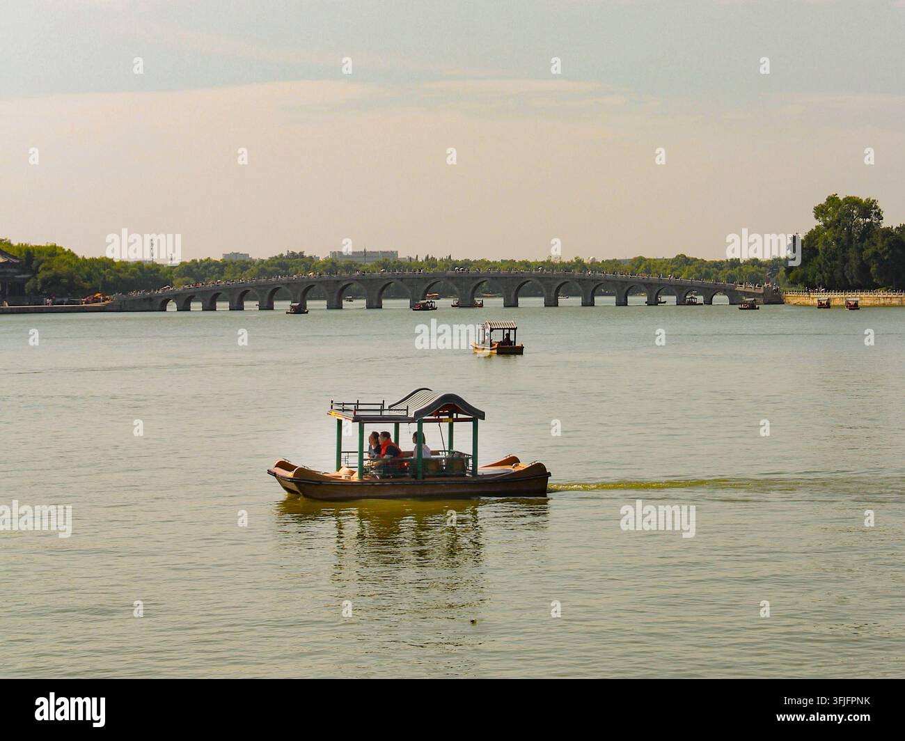Traditionelle hölzerne Touristenboote gleiten auf einem ruhigen Fluss mit einer langen historischen Steinbogenbrücke und Grünflächen im Hintergrund in China. Stockfoto