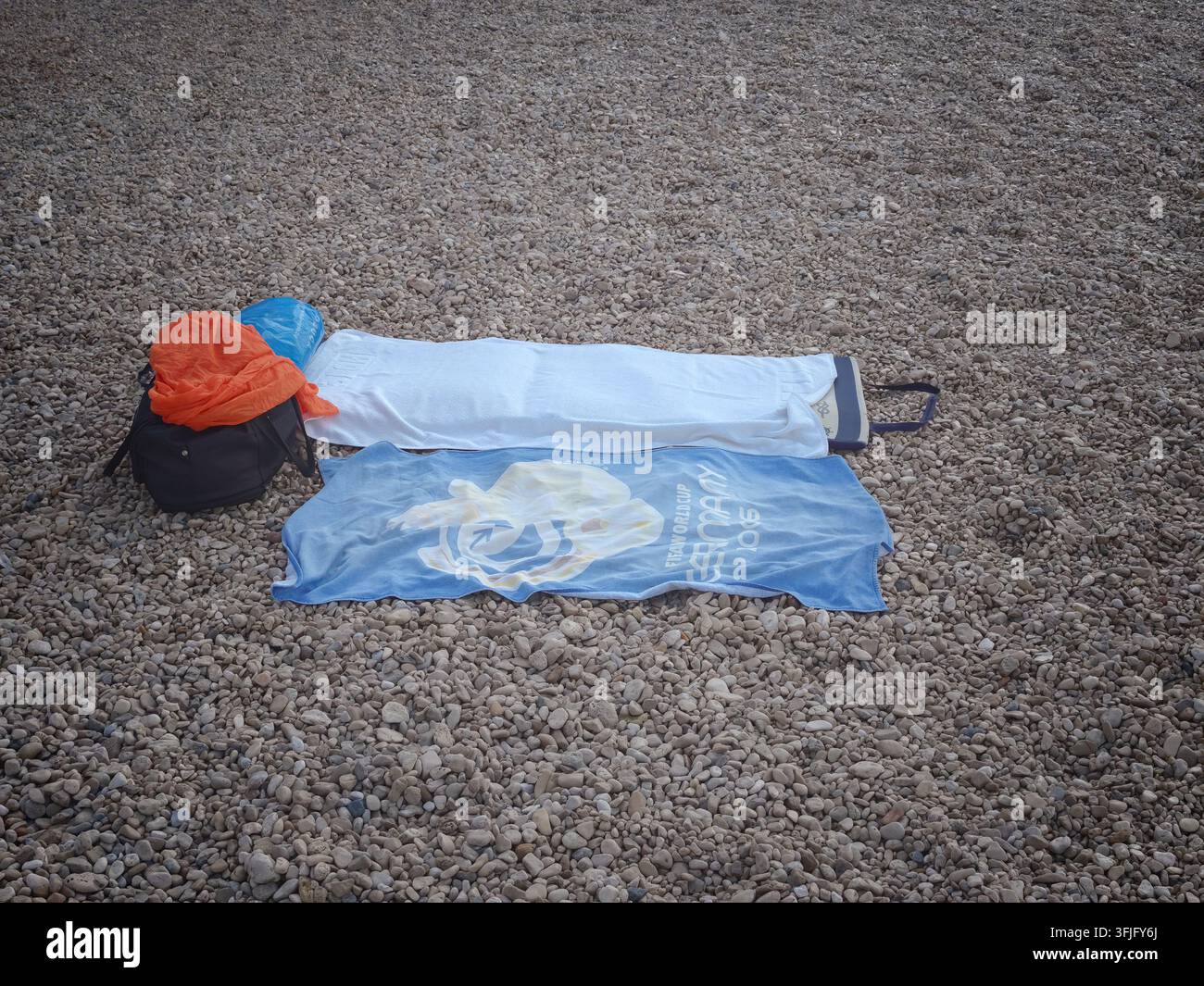 Eine schwarze Tasche, farbenfrohe Handtücher und Strandutensilien ordentlich auf einem felsigen Kieselufer angeordnet, das eine ruhige Sommerurlaubsstimmung einfängt. - Smartphone-aufgenommenes Stockfoto