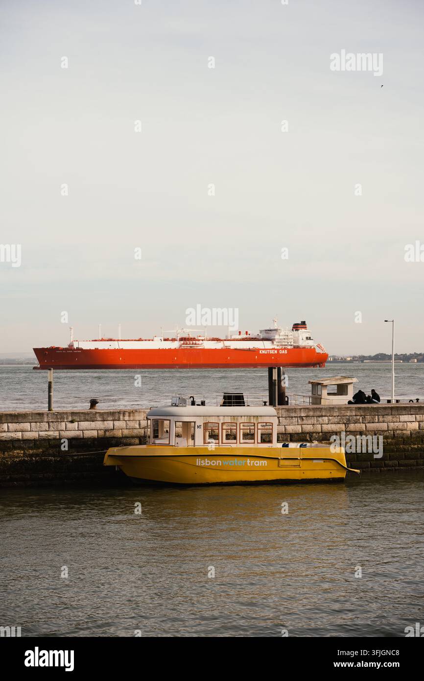 Lissabon, Portugal - 16. April 2026: Gelbes Passagierboot fährt neben der Uferpromenade, während ein rotes Tankerschiff unter dem Frühlingshimmel am Nachmittag auf dem Tejo vorbeifährt. Stockfoto