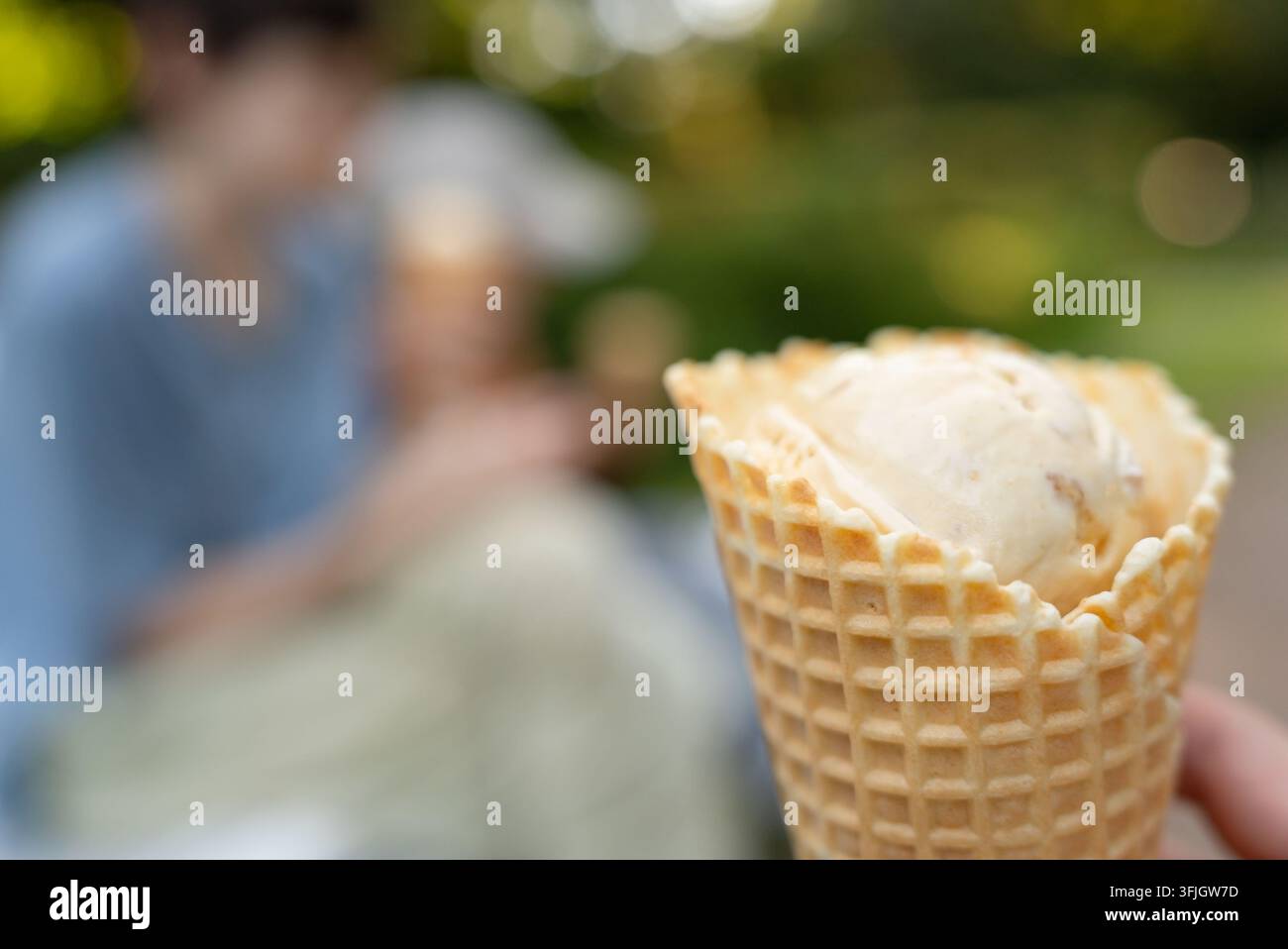 Eine verlockende knusprige Waffeltasche hält eine erfrischende Kugel Vanilleeis vor einem sonnigen Hintergrund im Freien. Dieser appetitliche Sommergenuss sieht bereit zum Essen aus in einer verschwommenen Parkumgebung. Stockfoto