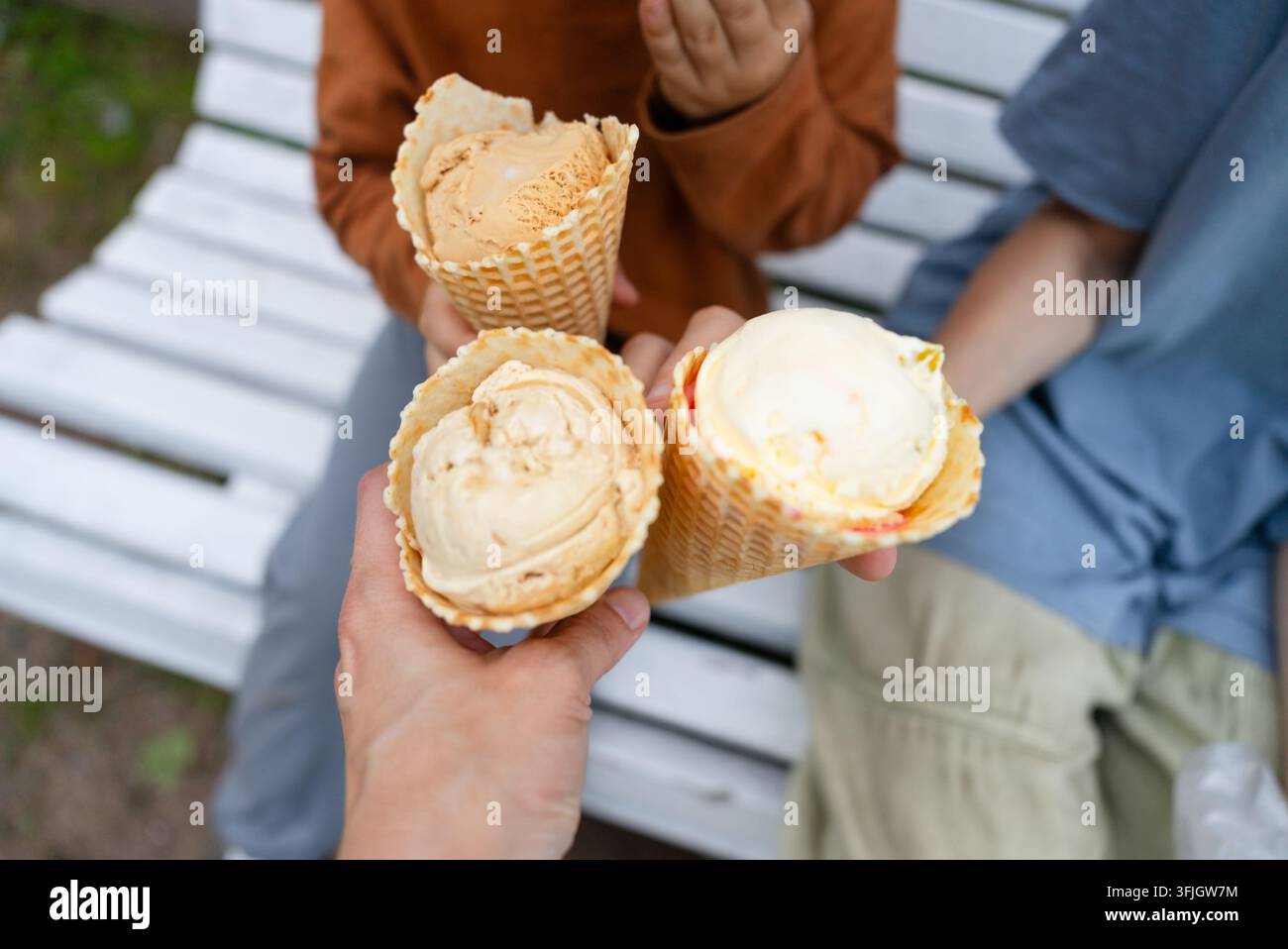 Eine Luftansicht zeigt einen Erwachsenen, der drei Eiswaffeln auf einer Parkbank hält, während Kinder in der Nähe spielen. Diese fröhliche Sommertreat-Szene unterstreicht die Freude am Teilen eines süßen Desserts im Freien. Stockfoto