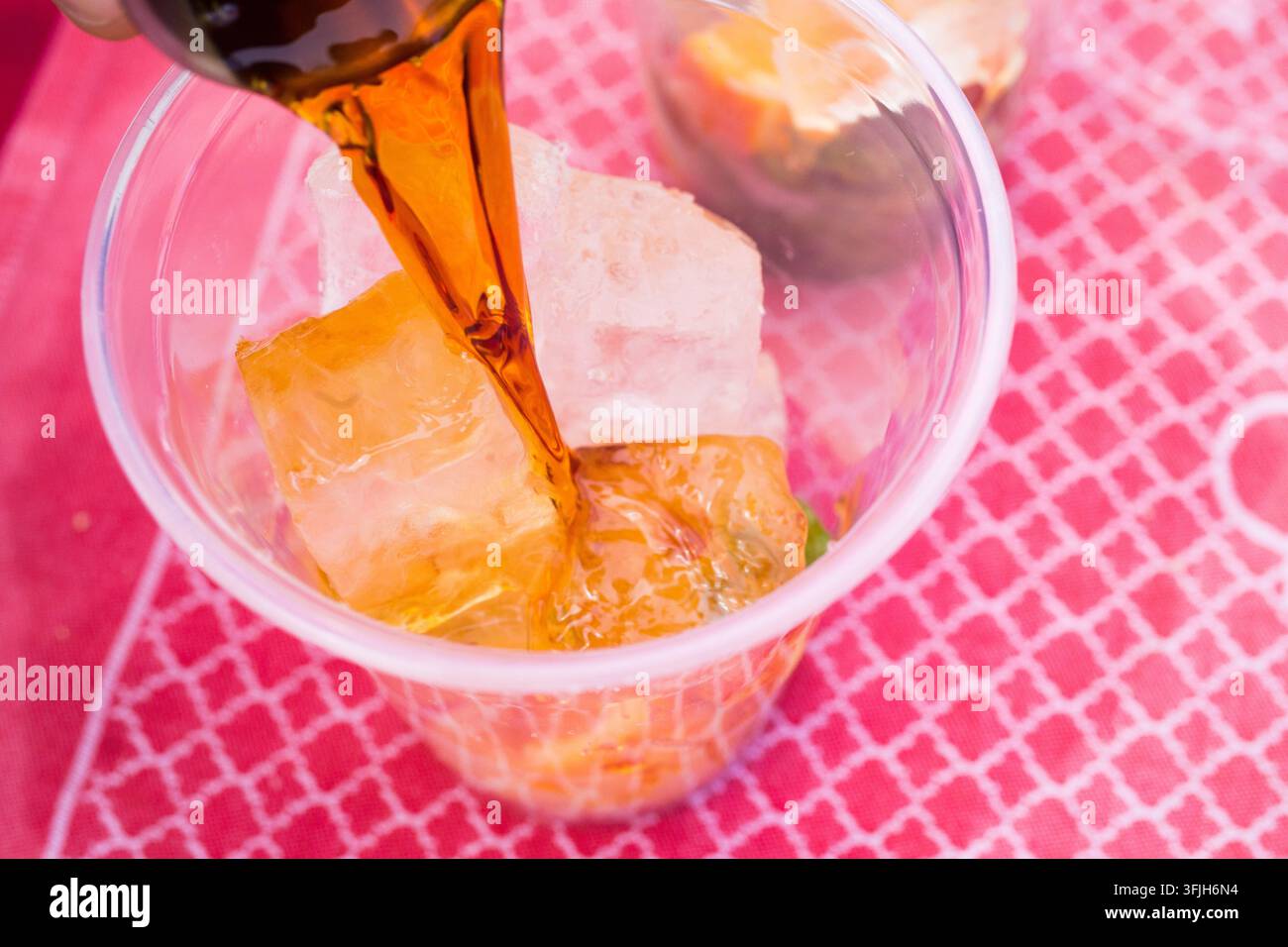Ein Straßenhändler gießt einen bernsteinfarbenen Cocktail über Eis in einen Plastikbecher auf einer rosa gemusterten Tischdecke, um ein erfrischendes Getränk zu servieren. Stockfoto