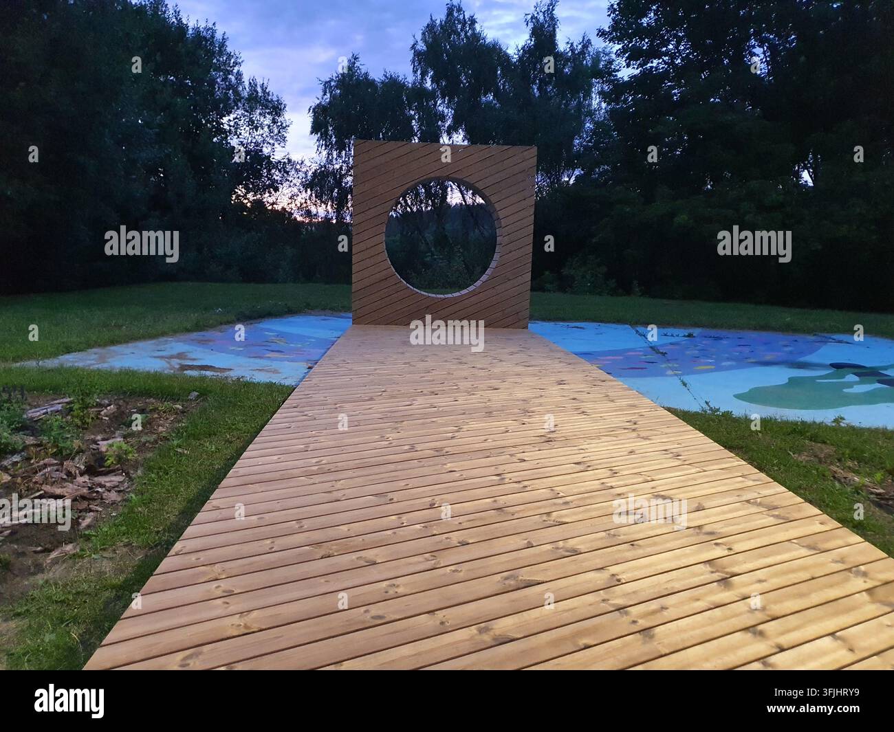 Ein Holzsteg führt zu einer modernen architektonischen Installation mit einer vertikalen Holzwand und einem großen runden Ausschnitt, der einem Portal ähnelt Stockfoto