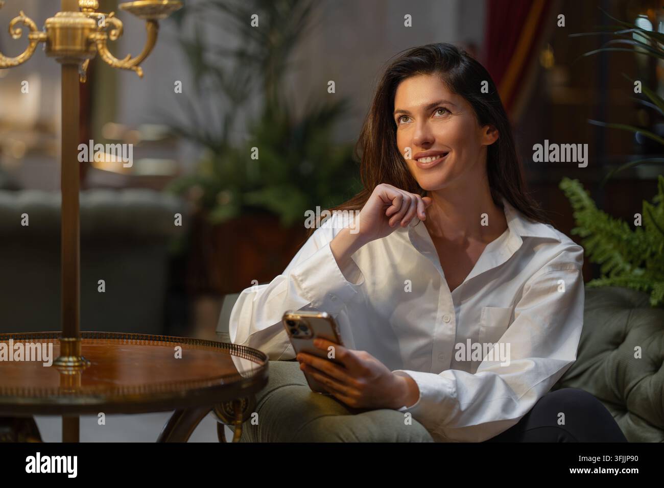 Porträt einer lächelnden jungen Frau, die ein Smartphone hält, während sie in einem stilvollen Interieur mit warmem natürlichem Licht sitzt. Stockfoto