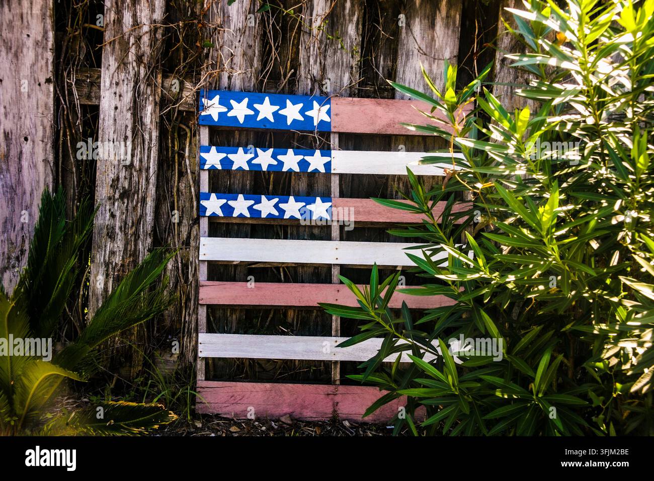 Eine mit blauen, weißen und roten Sternenmotiven bemalte Holzpalette, die der amerikanischen Flagge nachempfunden ist, lehnt an einem verwitterten Holzzaun. Stockfoto