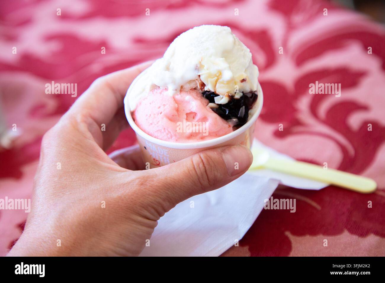 Hand, die eine kleine Tasse mit bunten Eisbechern mit cremiger Topping, dunkler Sauce und rosa Dessert hält, serviert über rotem gemustertem Tuch mit Serviette und Stockfoto