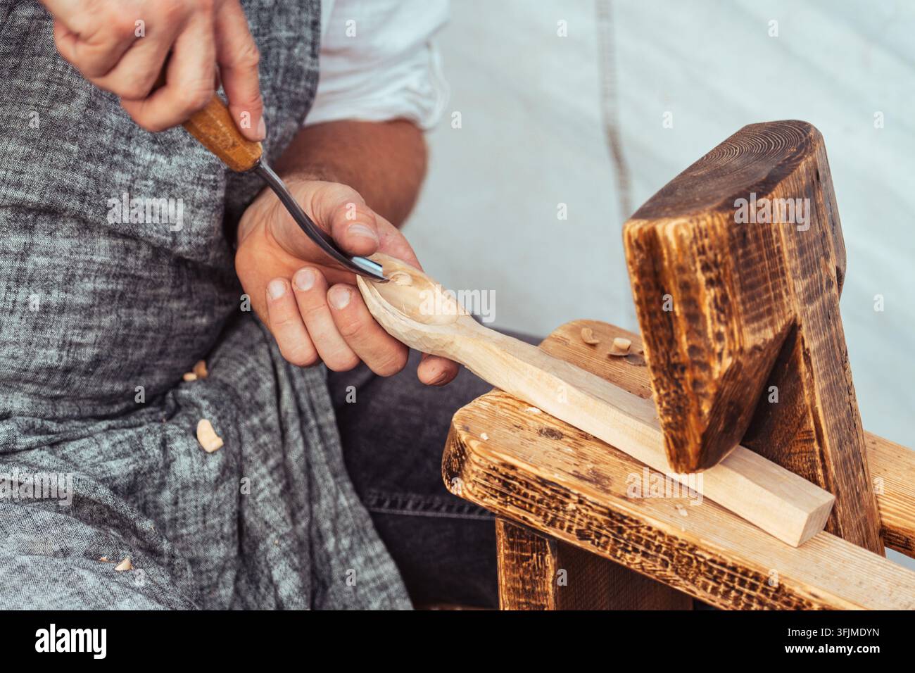 Nahaufnahme eines Handwerkers, der mit traditionellen Holzwerkzeugen einen Holzlöffel von Hand schnitzt. Handwerker in Schürze, die mit Holz arbeiten, Späne sichtbar Stockfoto