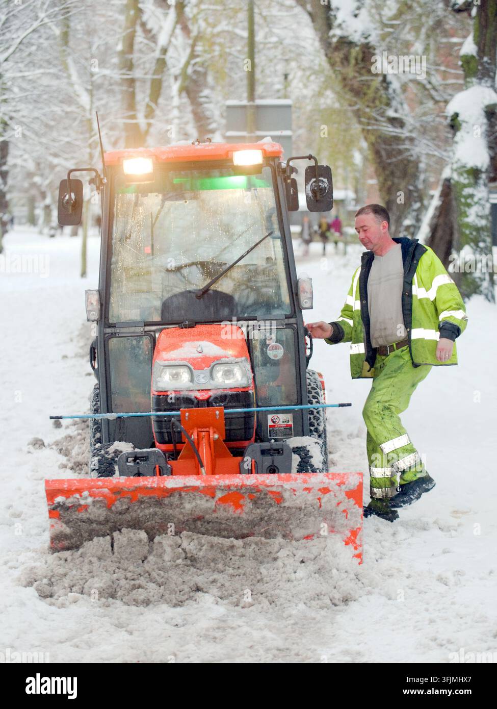 Ein Arbeiter, der eine Warnkleidung trägt, steigt aus einem kleinen Traktor mit Schneepflug während der Schneeräumungen im Winter. Stockfoto