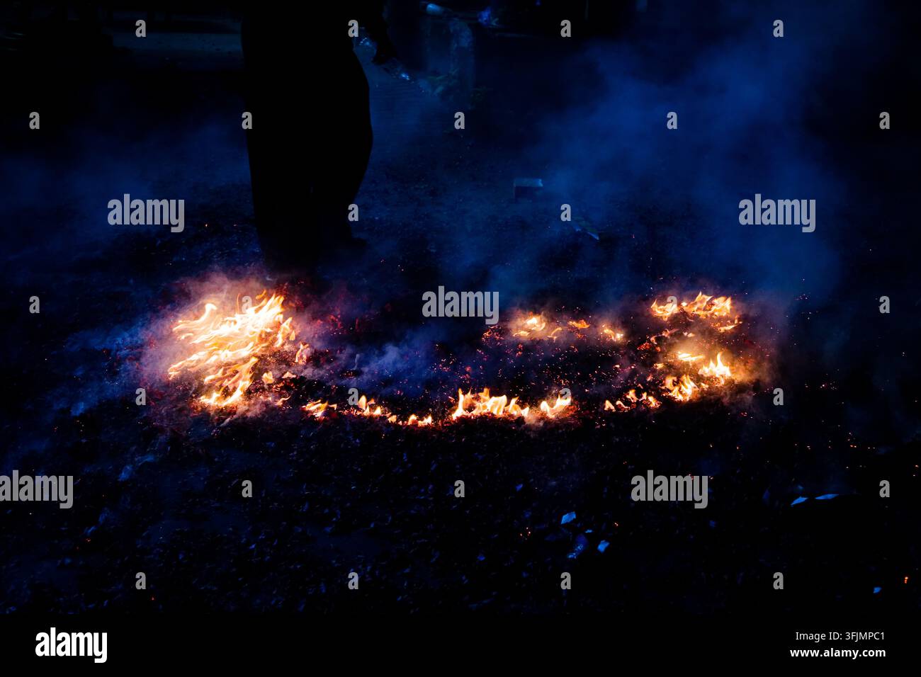 Eine silhouettierte Figur steht neben einem Ring aus glühenden Kohlen und Flammen auf dem Boden, Rauch zieht durch die tiefe blaue Nachtluft. Stockfoto
