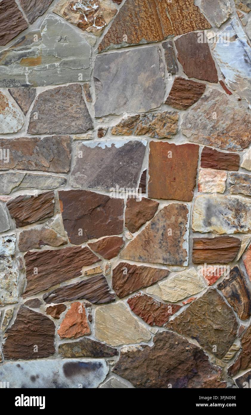 Mehrfarbige maschinell geschnittene Furnierstein- und vertiefte Mörtelwandoberfläche, die kontrastierende glatte und stark texturierte Steine in einer vertikalen Komposition zeigt Stockfoto