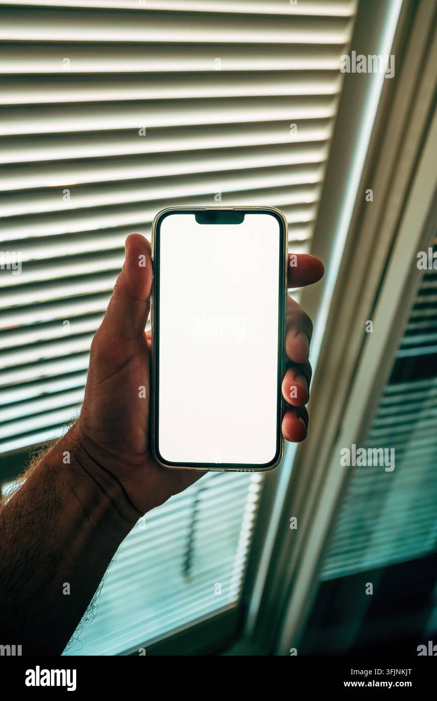Hand hält ein modernes Smartphone mit einem leeren weißen Bildschirm-Mockup vor Jalousien mit sanftem Tageslicht. Selektive Fokussierung. Stockfoto