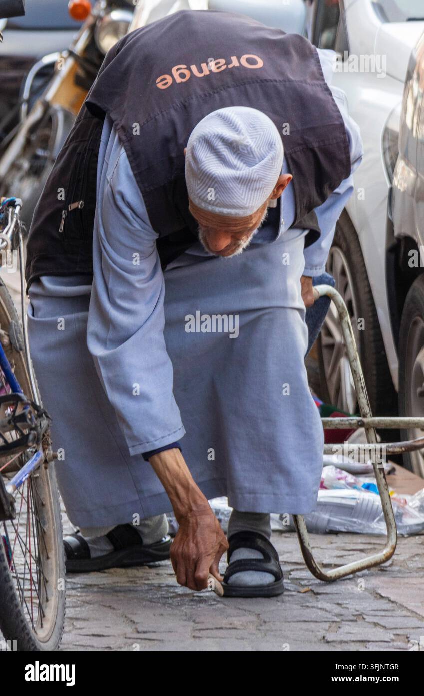 Älterer Mann repariert Sandale mit einem Stein. Ein Anwohner, der eine Orange-Marke-Weste trägt, beugt sich auf dem Gehweg in Marrakesh hinunter. Stockfoto