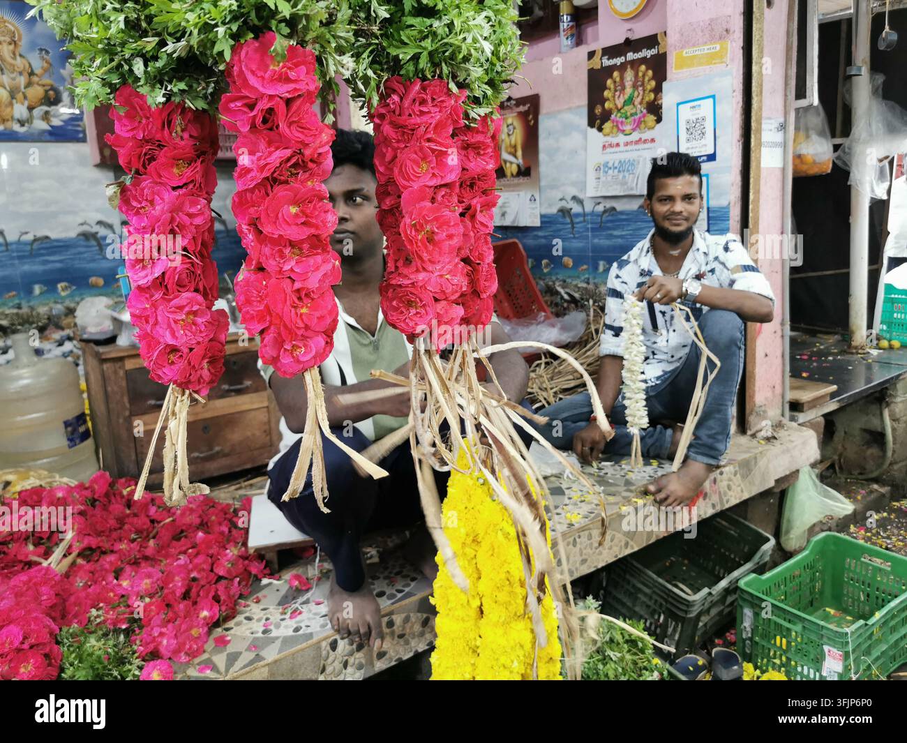 PUDUCHERRY, INDIEN - 18. JANUAR 2026: Zwei Blumenhändler sitzen hinter hängenden Girlanden aus Rosen und Ringelblumen an einem Blumenstand auf einem Markt. Stockfoto