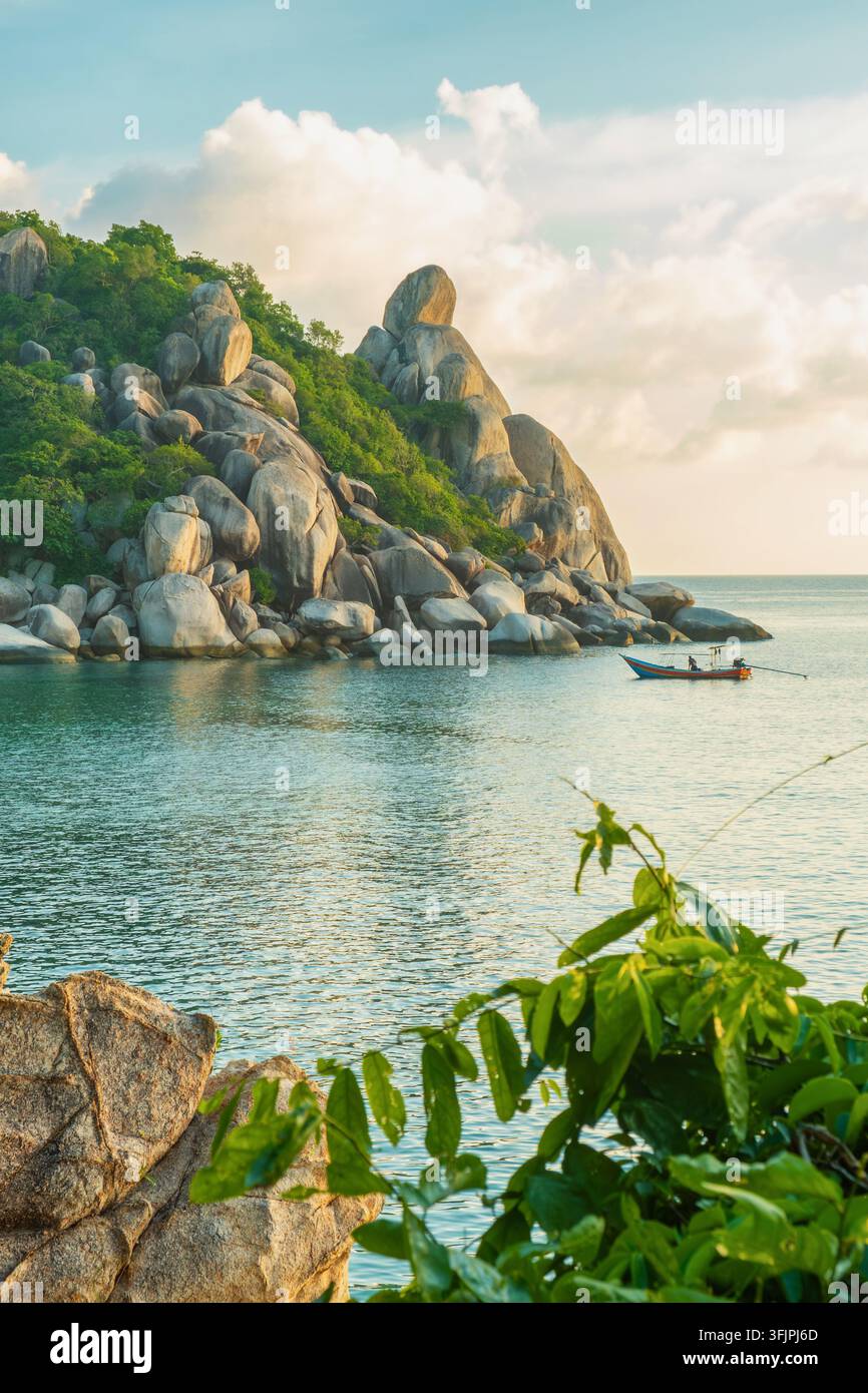 Granitblöcke und tropische Vegetation in der Nähe von Freedom Beach auf Koh Tao, Thailand, mit traditionellem Boot auf ruhigem Wasser bei Sonnenuntergang. Inselreise Stockfoto