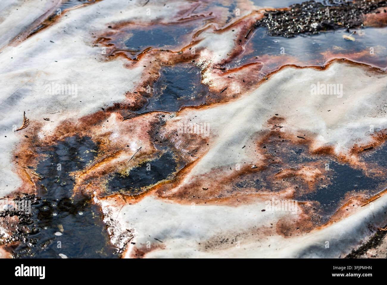 Eine Hochwinkelaufnahme zeigt schmutziges Wasser und Rostflecken, die sich über ein weißes Textiltuch im Freien ausbreiten. Pfützen bilden dunkle Flecken auf dem Stoff. Stockfoto