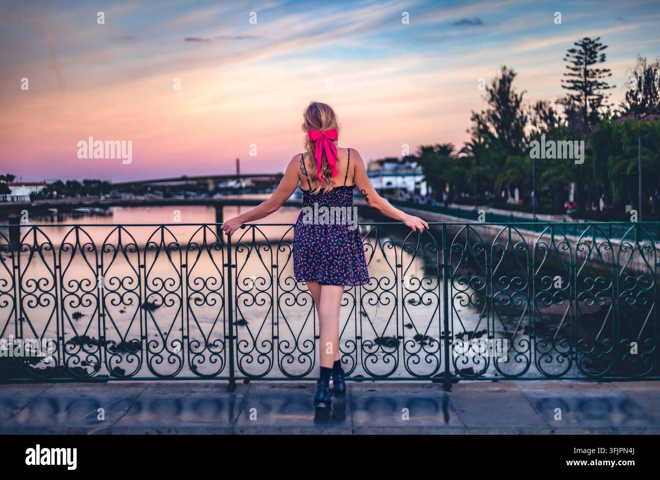 Junge Frau mit einer rosa Schleife im Haar, die sich an einem Flussbrückengeländer bei Sonnenuntergang in Tavira, Algarve, Portugal, lehnt. Stockfoto