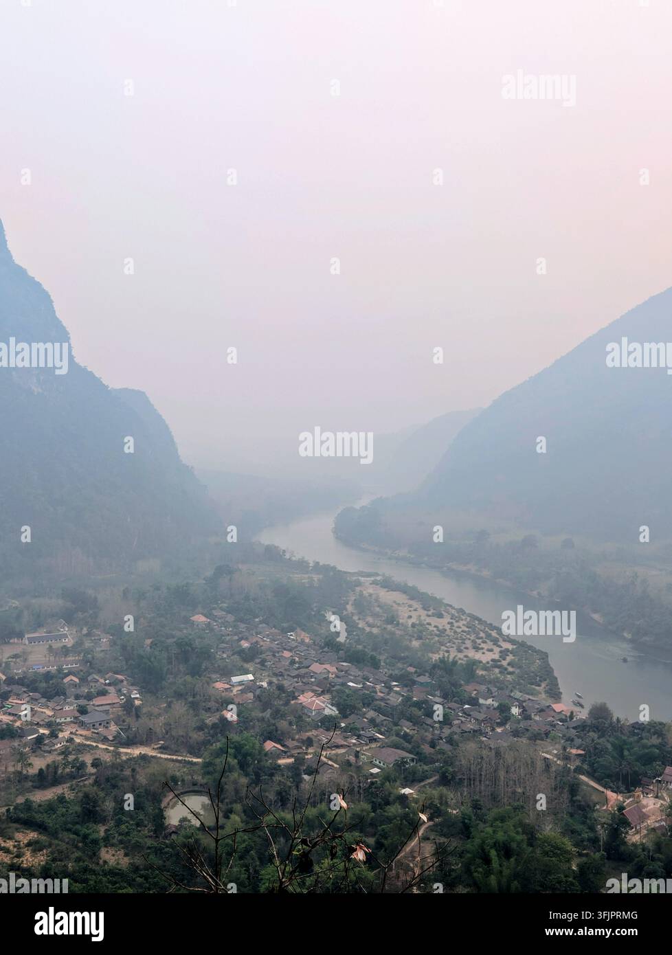 Hochwinkel-Landschaftsfotografie eines Flusses, der sich durch ein kleines ländliches Dorf schlängelt, umgeben von steilen Kalkstein-Karstbergen in dichtem Dunst. - Smartphone-aufgenommenes Stockfoto