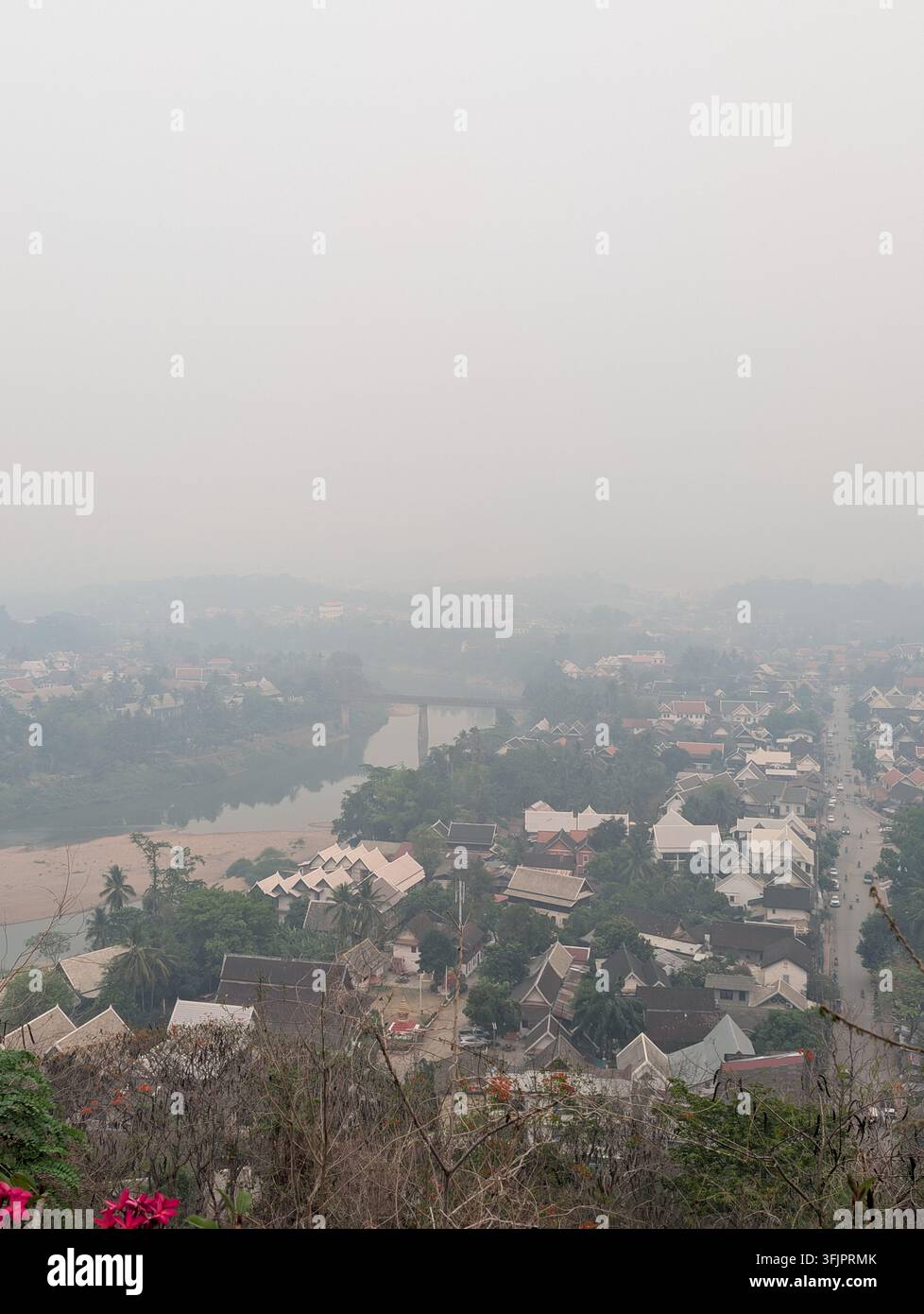 Hohe Blickwinkelansicht mit Blick auf traditionelle asiatische Dächer, einen gewundenen Fluss und Brücken in Luang Prabang unter einem nebligen, dunstigen Himmel, aufgenommen vom Aussichtspunkt des Berges Phousi - Smartphone-aufgenommenes Stockfoto