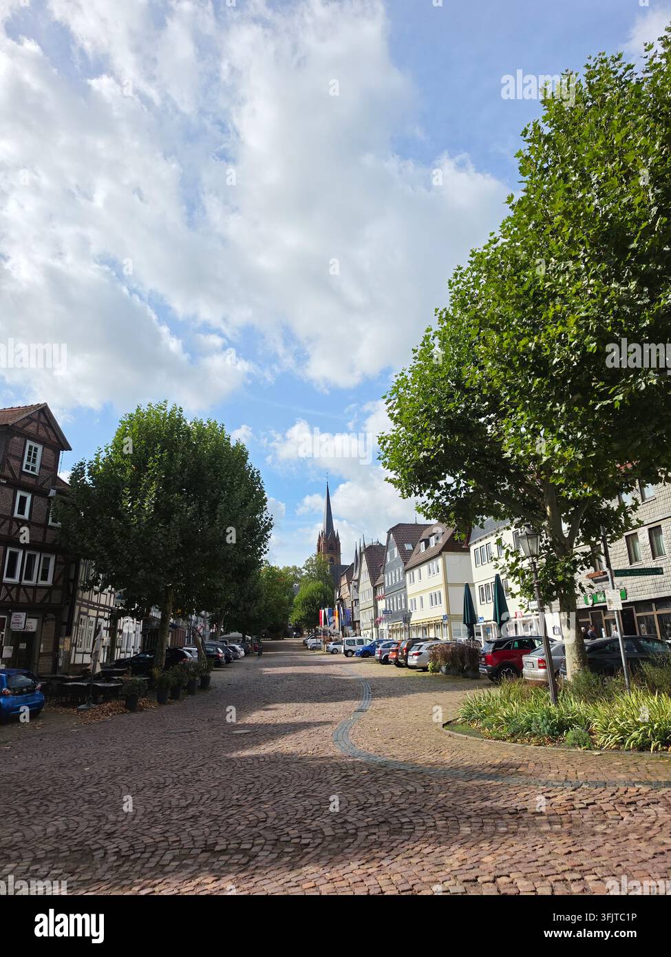 Ein weiter Blick auf eine historische europäische Kopfsteinpflasterstraße, gesäumt von grünen Bäumen, traditionellen Gebäuden und einem Kirchturm in der Ferne unter einer Wolke - Smartphone-aufgenommenes Stockfoto