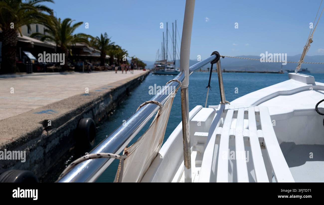POV-Nahaufnahme vom Deck eines weißen Holz-Vergnügungsbootes, das entlang einer sonnigen Resort-Promenade mit Palmen und Café-Terrassen festgemacht ist. Ideal für den Sommer v Stockfoto