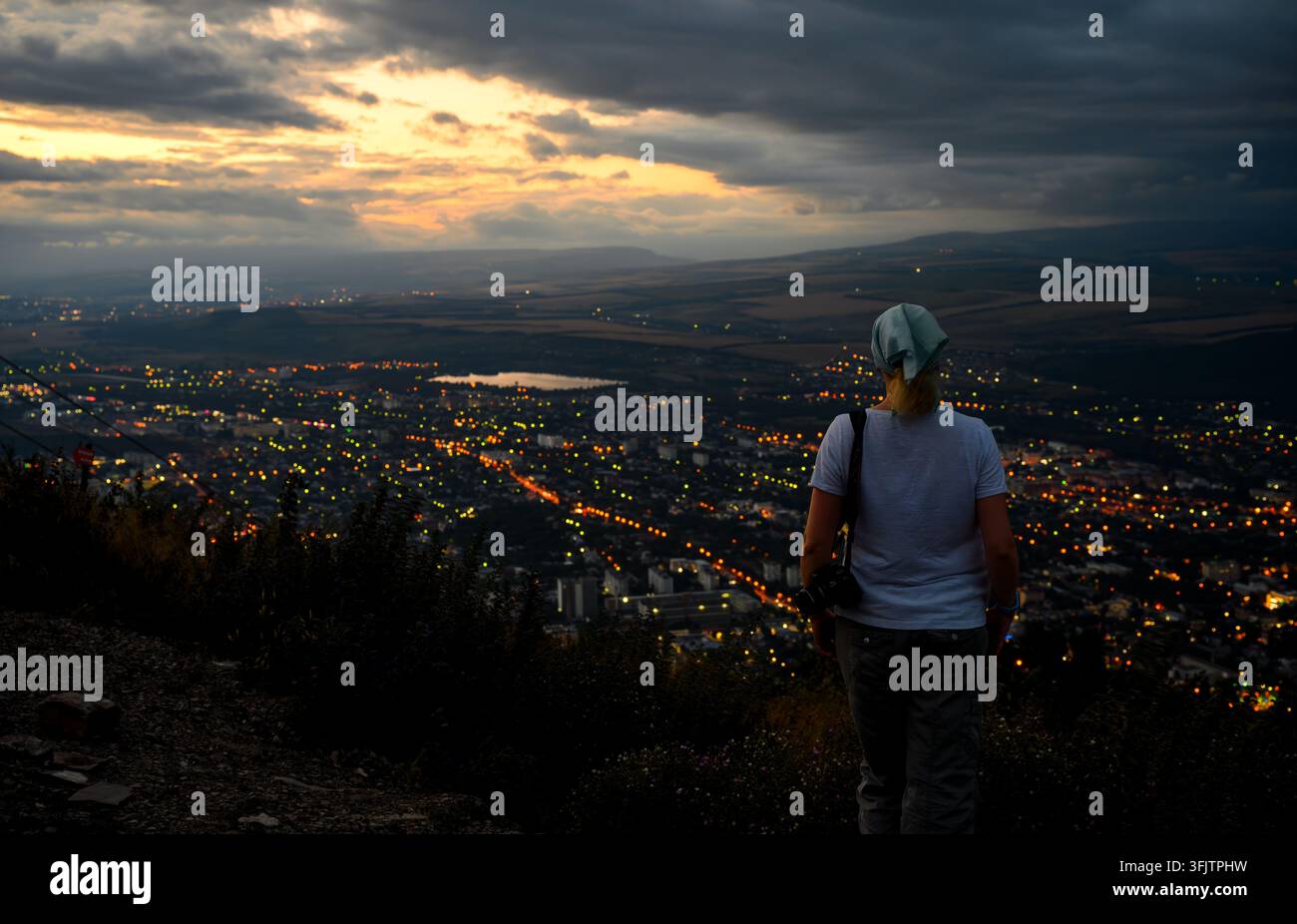 Eine Rückansicht einer Fotografin, die auf einem hohen Aussichtspunkt steht und bei Dämmerung über eine weitläufige Stadt blickt. Sie trägt ein weißes T-Shirt, grau Stockfoto