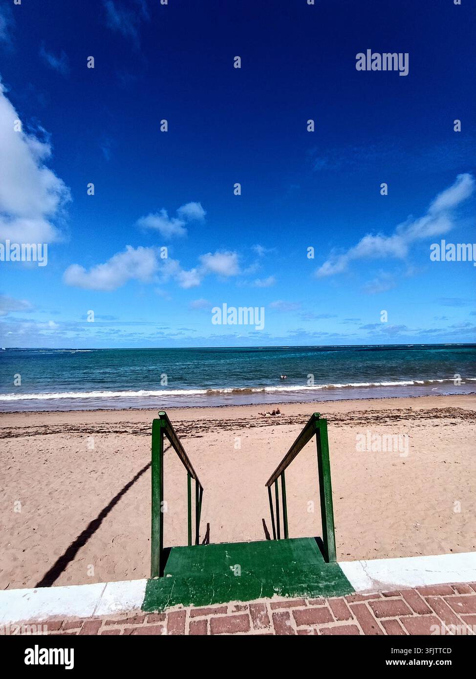 Grün geländerte Stufen führen von einer Uferpromenade auf den sandigen Strand von Maragogi Beach, Alagoas, Brasilien, unter einem strahlend blauen Himmel. Stockfoto