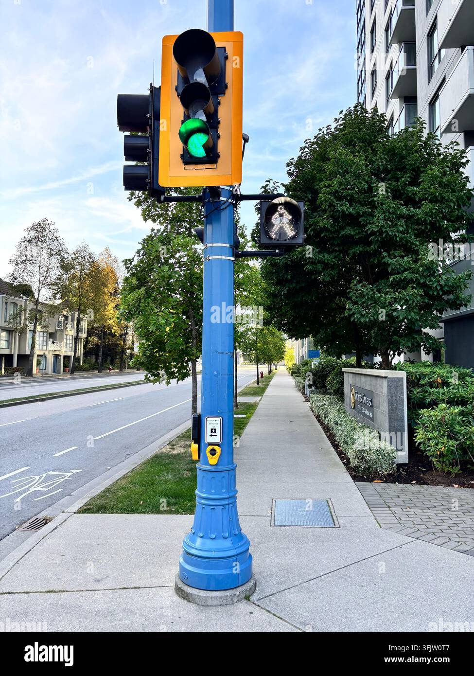 Eine hellblaue Ampelmast steht auf einem Gehweg mit dem grün leuchtenden Signal und leitet Fahrzeuge und Fußgänger sicher durch die Stadtstraße. - Smartphone-aufgenommenes Stockfoto