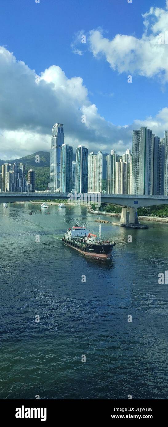 HONGKONG - 30. AUGUST 2025: Ein Handelsschiff fährt durch ein städtisches Wasserstraßensystem unter einer modernen Brücke in Hongkong. Stockfoto