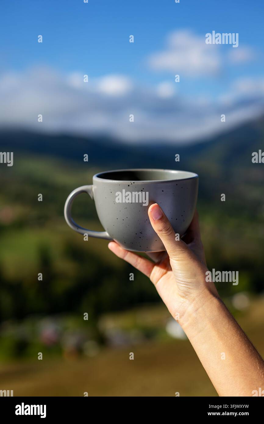 Hand hält eine Keramiktasse vor einer malerischen Bergkulisse. Die Hand hält eine graue Keramiktasse im Sonnenlicht mit einem verschwommenen grünen Bergtal, Wolken und Stockfoto