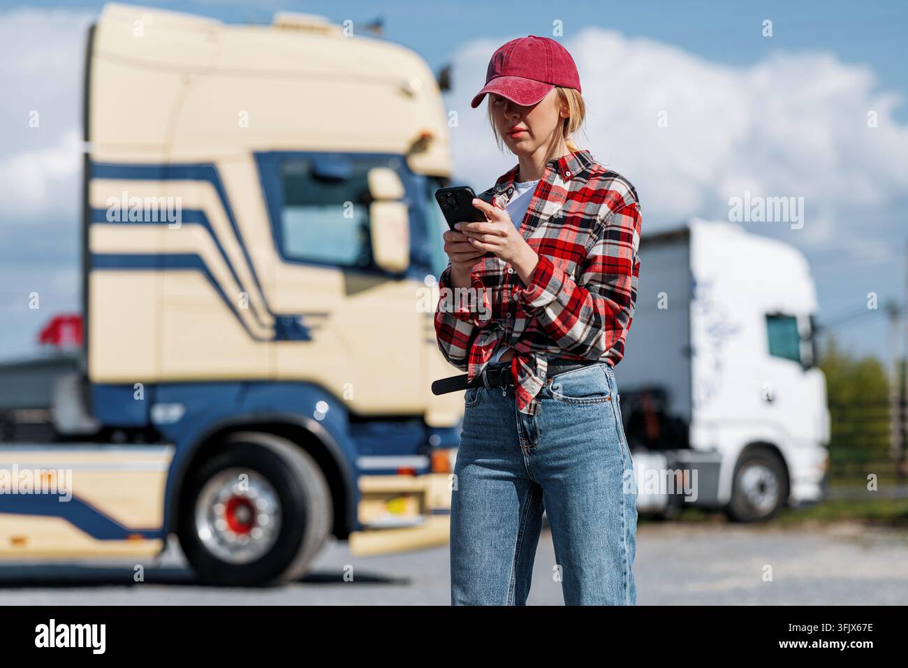 Eine Truckerin in einem karierten Hemd und Baseballmütze nutzt ihr Smartphone im Freien in der Nähe von zwei großen Sattelzügen an einem sonnigen Tag Stockfoto