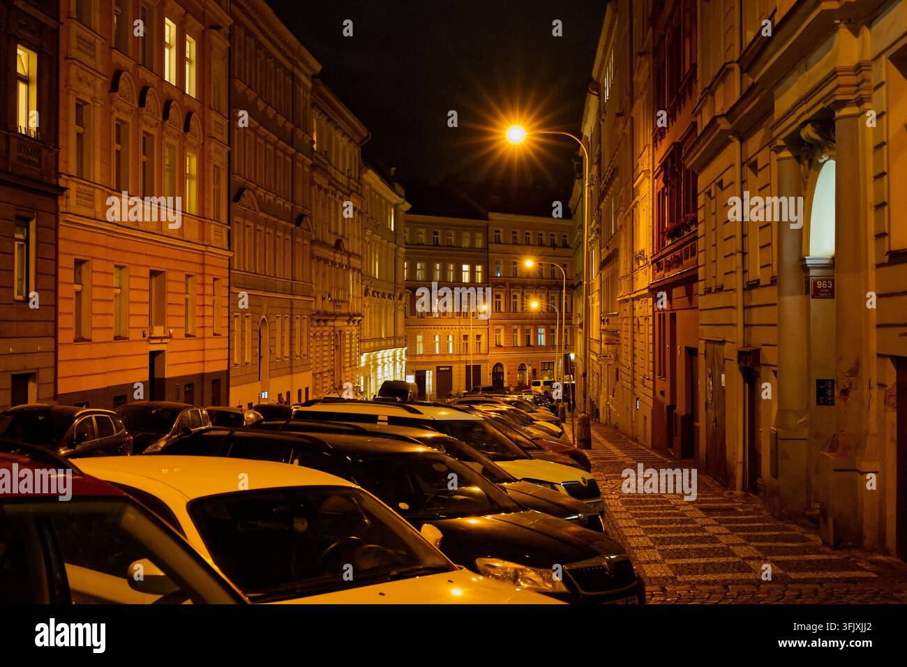 Abhängige Stadtstraße voller geparkter Autos und historischer Wohnhäuser unter warmen Straßenlaternen bei Nacht. Stockfoto