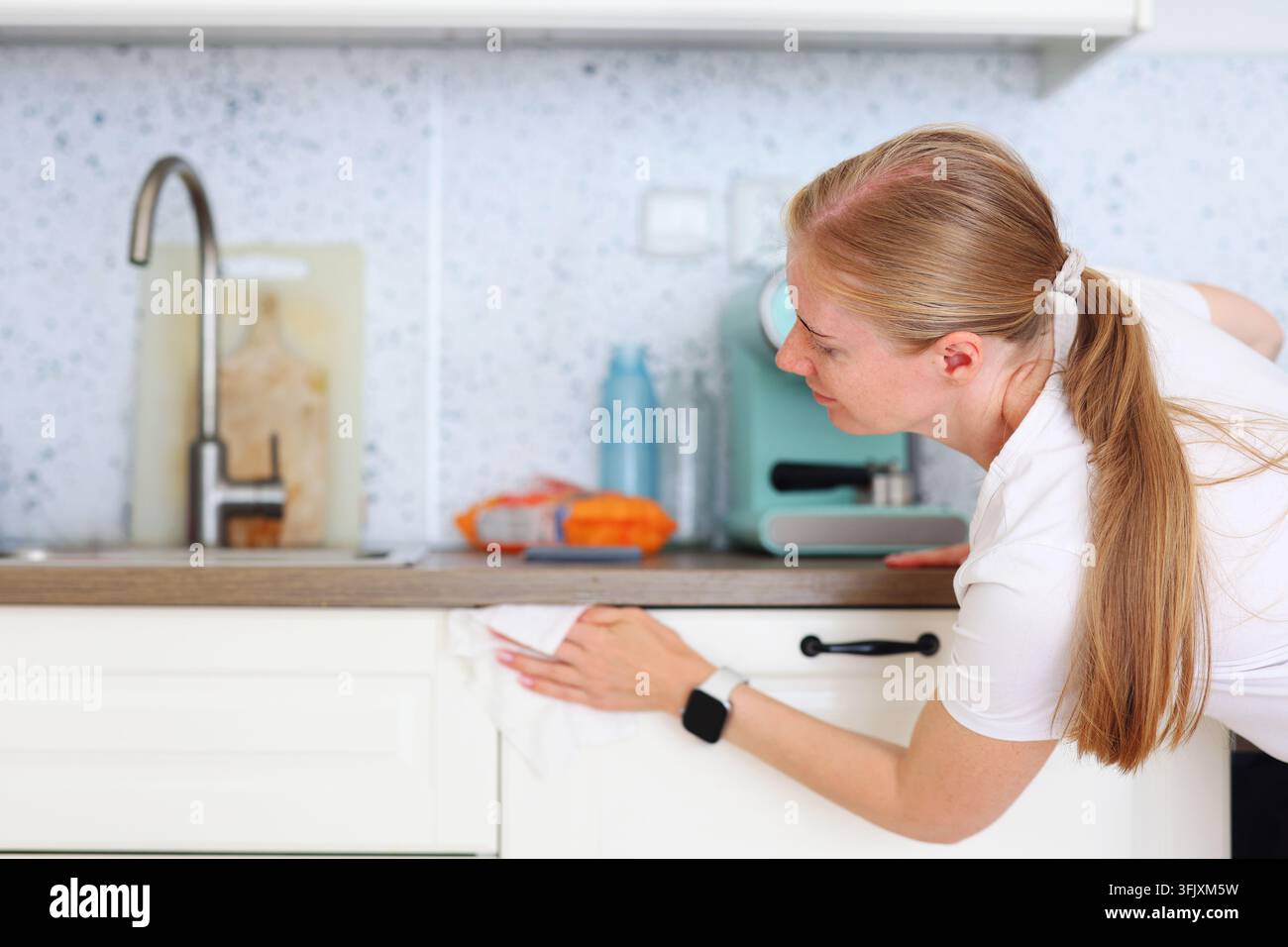 Lächelnde Frau wischt Küchenschränke mit einem Tuch ab, repräsentiert alltägliche Hausarbeit, Sauberkeit und ein gesundes Zuhause. Stockfoto