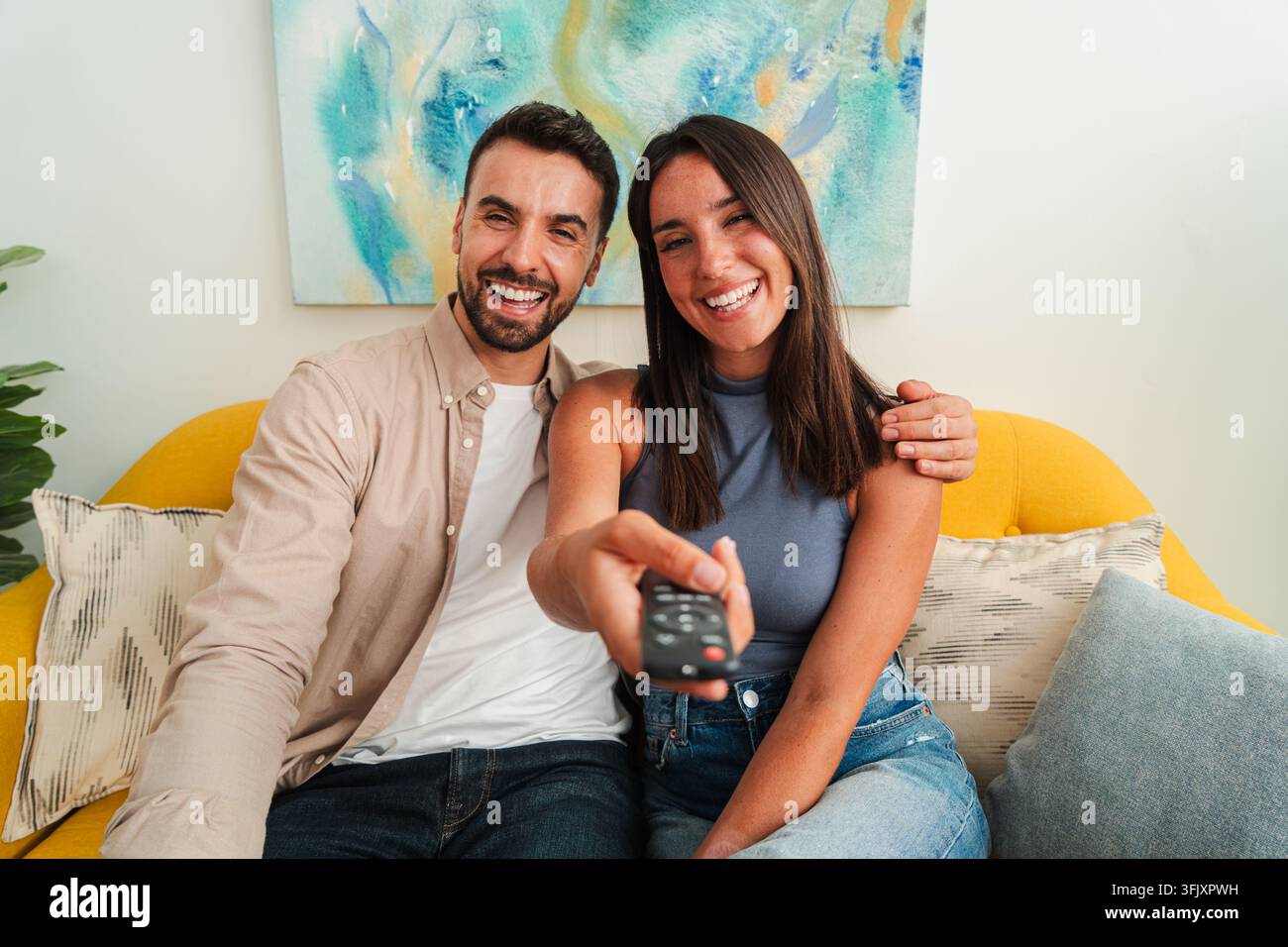 Ein warmes Porträt zeigt ein lächelndes Paar auf einem hellgelben Sofa, einer zeigt eine TV-Fernbedienung in Richtung des Betrachters, während sie einen entspannten, intimen Moment teilen Stockfoto