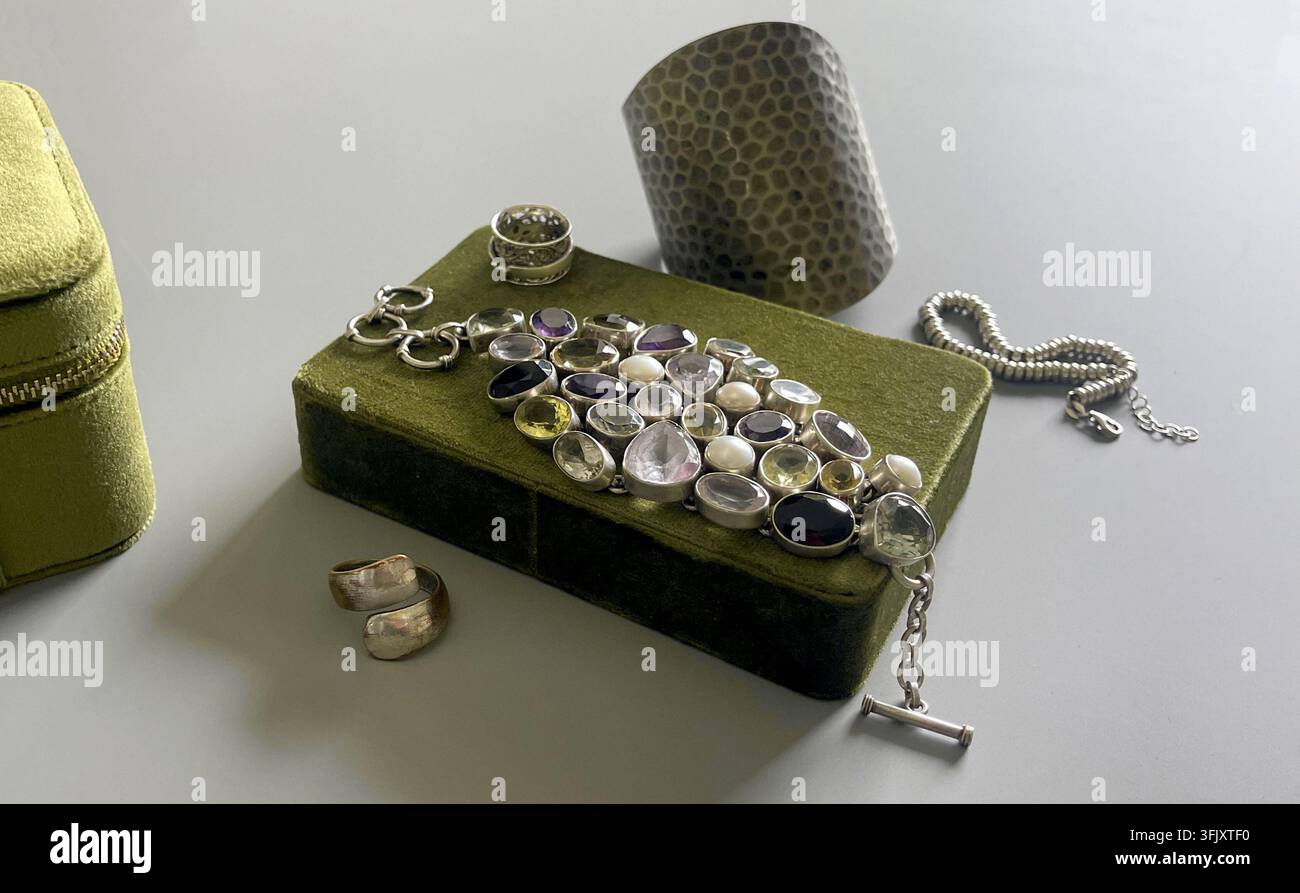 Ein Satz silberner Schmuckstücke ist auf einer grünen Schachtel ausgelegt. Die Kollektion umfasst Ringe, ein Armband und eine Manschette. Sonnenlicht strömt durch die nahe gelegene w Stockfoto