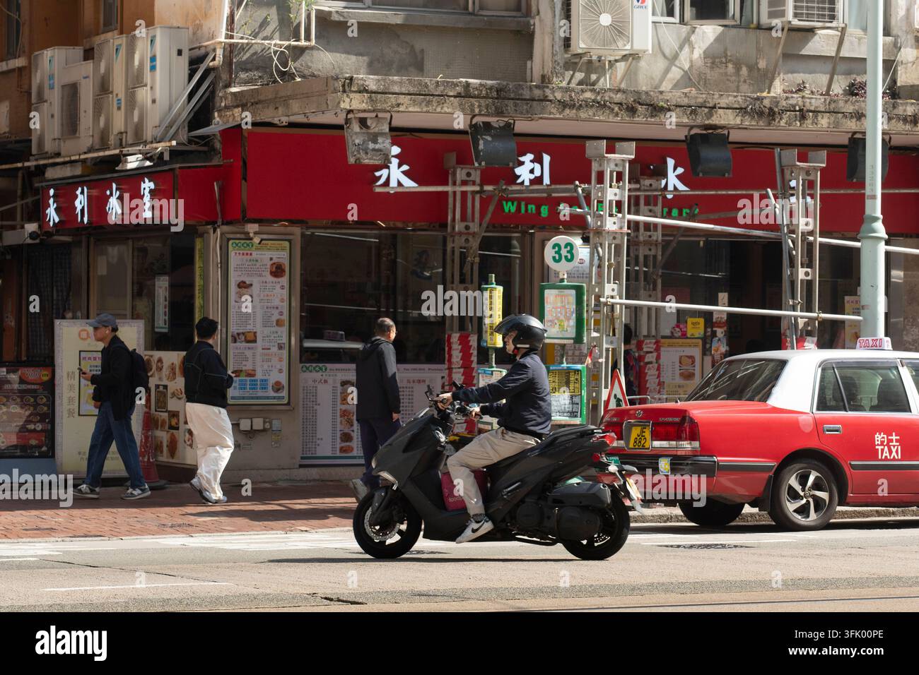 Ein Kurier fährt mit einem Roller am Wing Lee Restaurant in North Point, Hongkong, vorbei und navigiert durch den Straßenverkehr, um Essen zu liefern. Stockfoto
