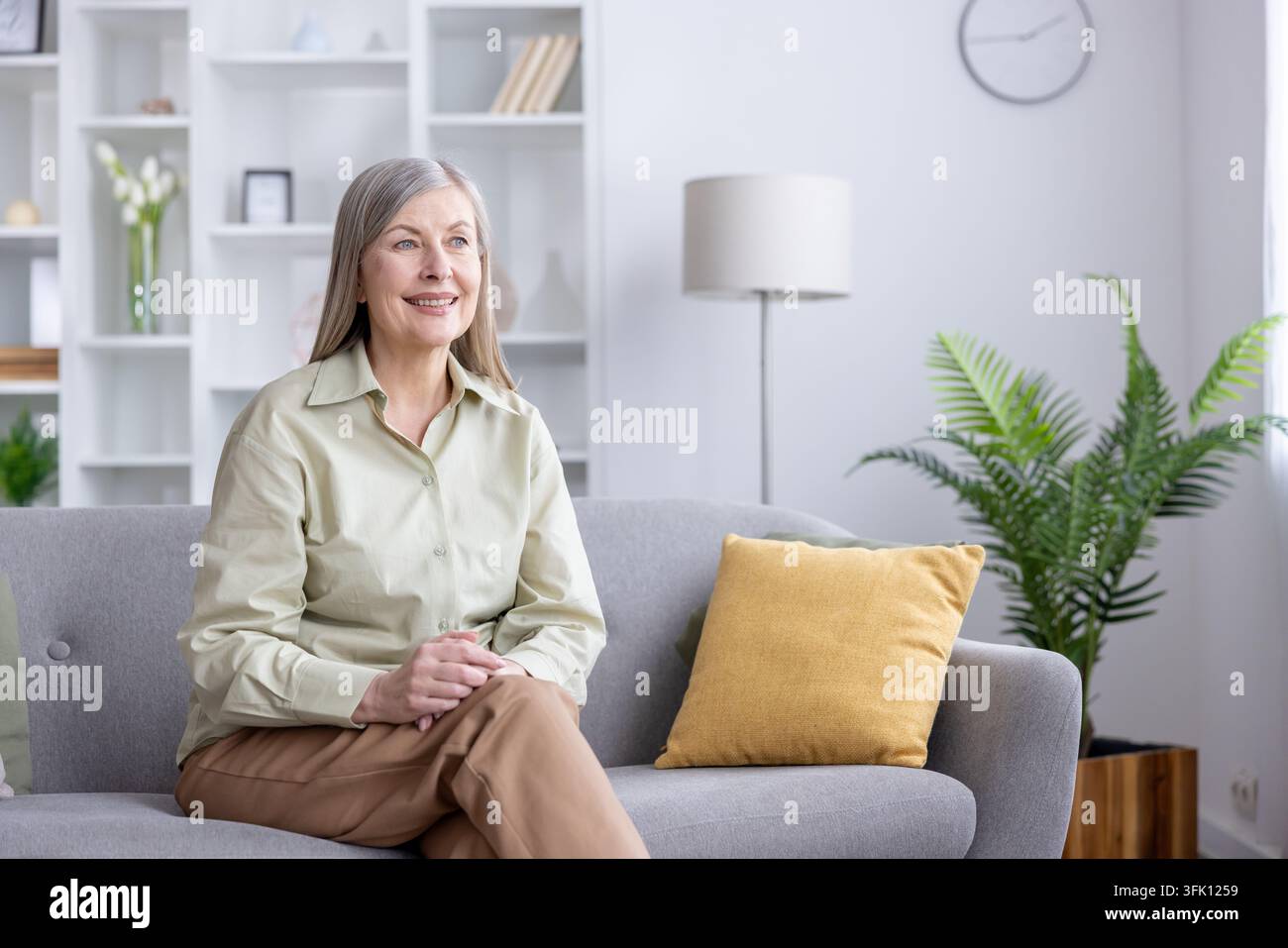 Seniorin sitzt bequem auf einer grauen Couch in einem hellen Wohnzimmer, lächelt friedlich und genießt einen ruhigen Moment der Entspannung und Zufriedenheit zu Hause Stockfoto