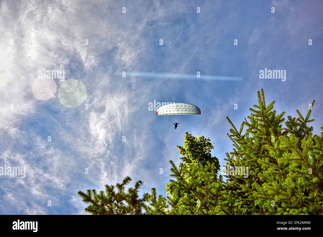Niedrigwinkelaufnahme eines weißen Gleitschirmfliegers, der über einen grünen Baum unter einem blauen Himmel mit leichten Wolken in Savoie, Frankreich, fliegt und Outdoor-Abenteuer einfängt Stockfoto