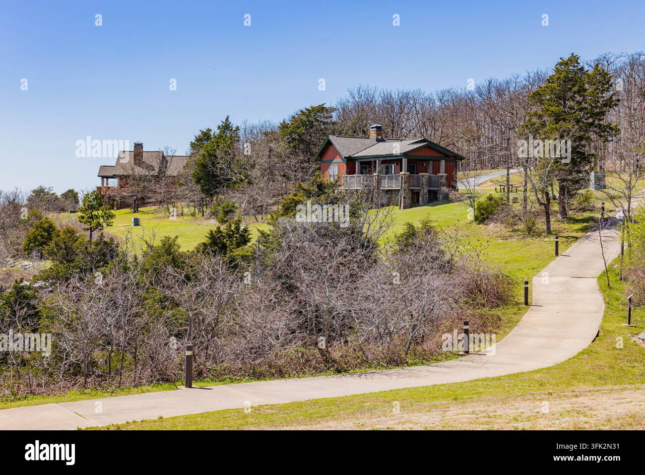 Zwei rustikale Holzhäuser überblicken einen gewundenen Betonweg durch eine hügelige Landschaft voller Bäume und grünem Gras. Stockfoto