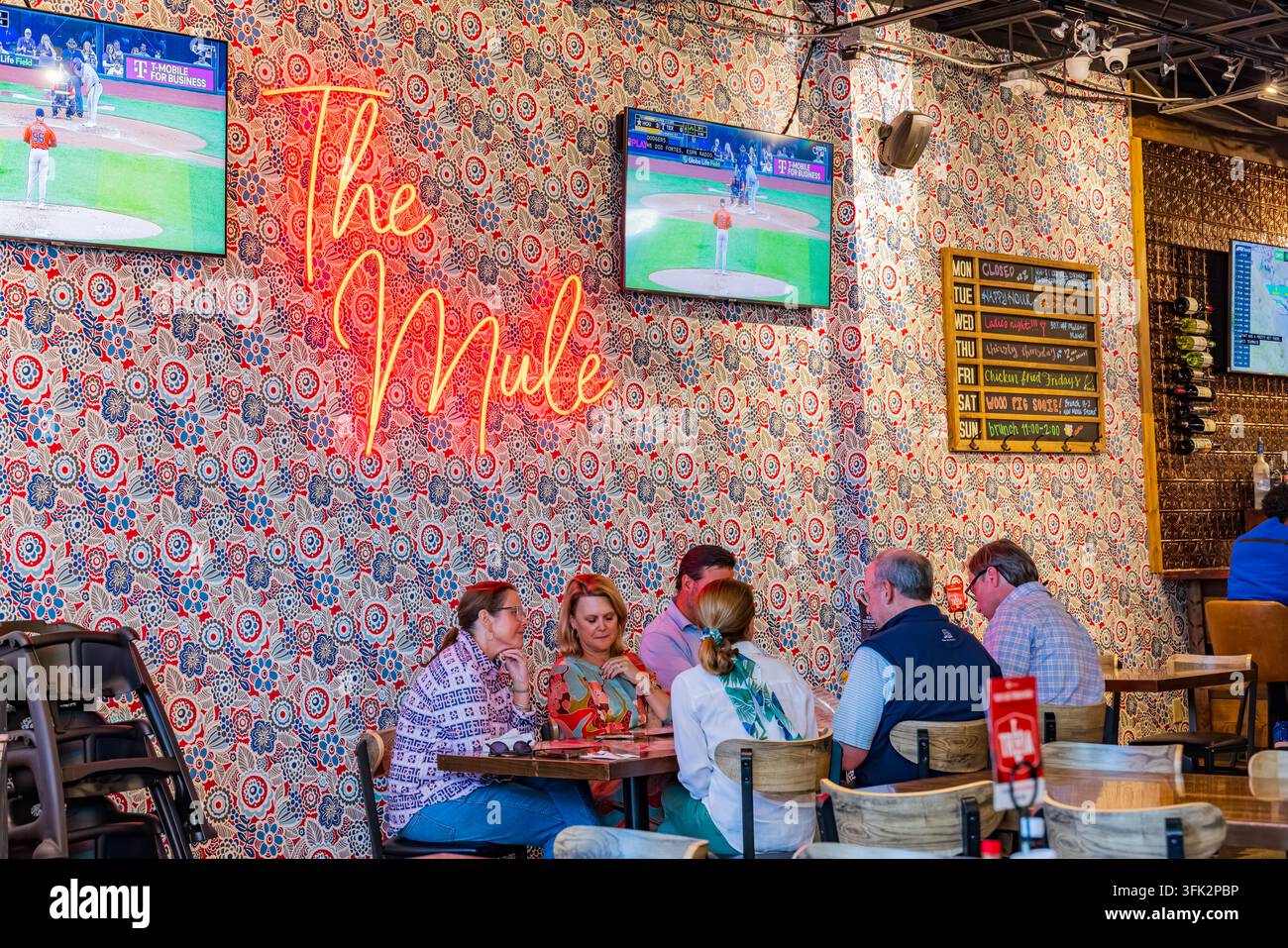 Arkansas, Vereinigte Staaten, 07.04.2024 – Freunde versammeln sich um einen Holztisch in einem farbenfrohen Restaurant mit gemusterter Tapete und einem leuchtenden Neonlicht Stockfoto