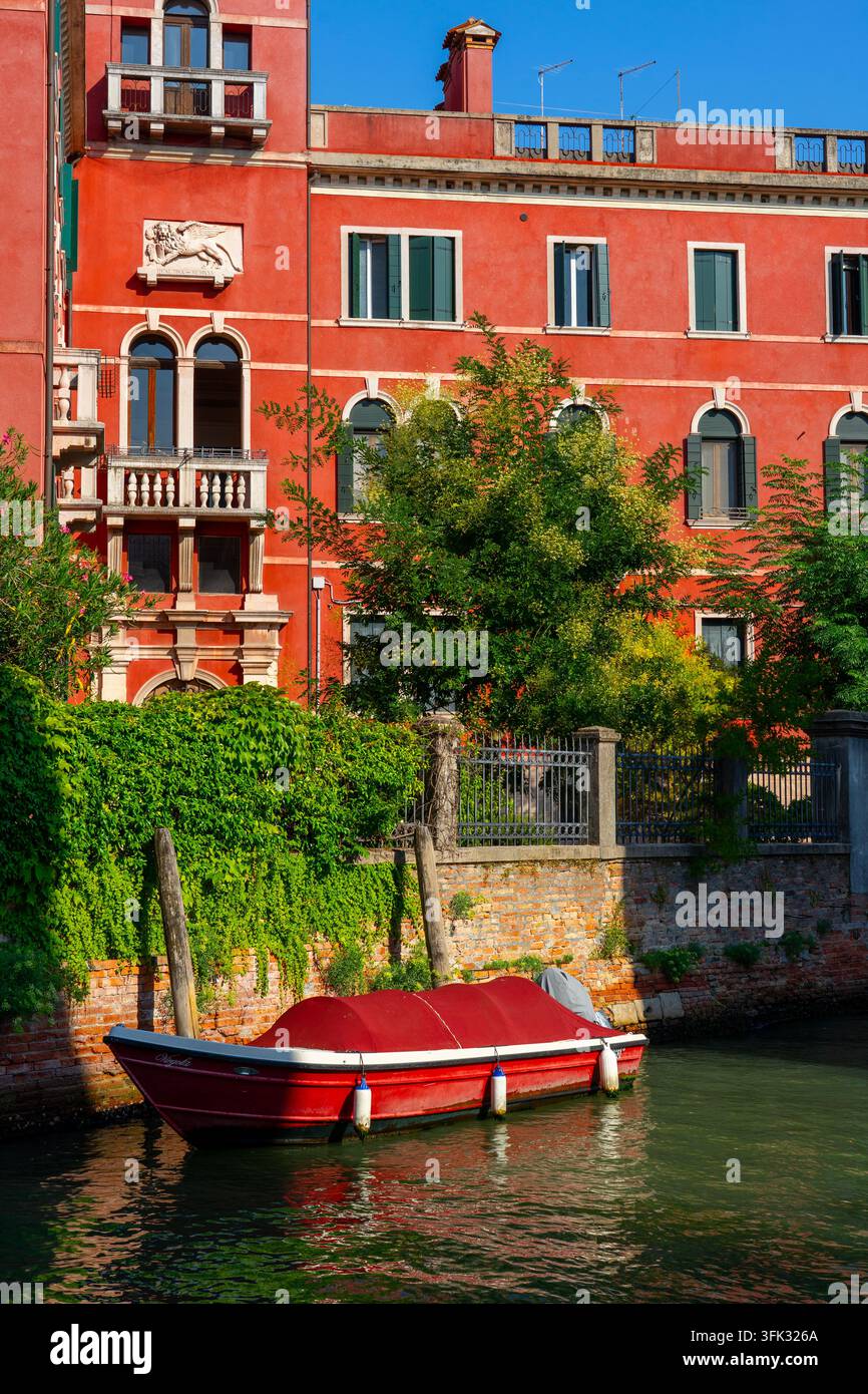Rotes venezianisches Palais und ein festgemachtes Boot entlang eines ruhigen Kanals in Venedig . Stockfoto