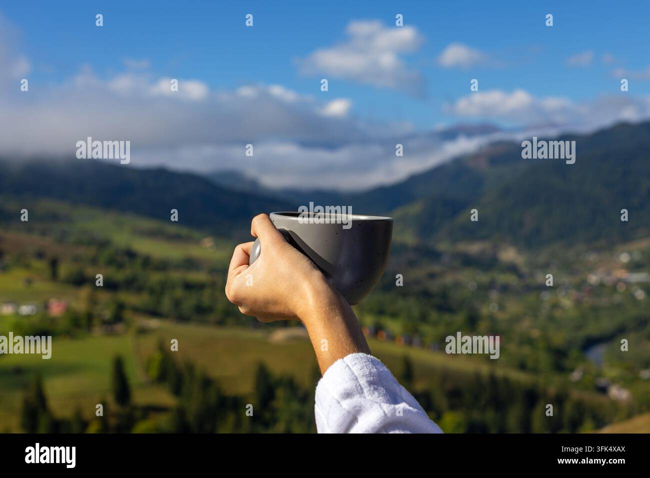 Hand hält eine Keramiktasse über ein malerisches Bergtal. Eine Hand in einem weißen Bademantel hält eine Keramiktasse im Sonnenlicht vor einem verschwommenen grünen Bergtal Stockfoto