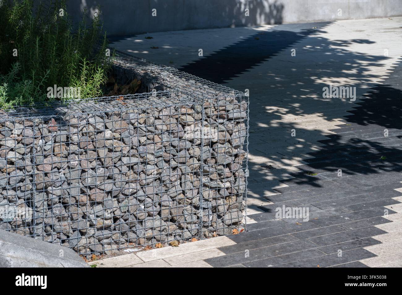 Eine moderne mit Steinen gefüllte Drahtkorb-Stützmauer grenzt an eine sonnenbeschienene gepflasterte Terrasse mit üppigen grünen Pflanzen und gesprenkelten Baumschatten Stockfoto